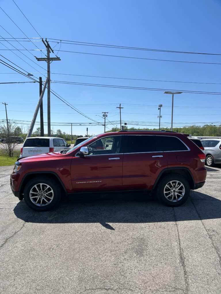 Red Jeep Grand Cherokee parked in a lot with power lines and poles overhead against a clear blue sky.