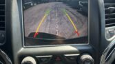In-car touchscreen showing a rearview camera view with green and yellow guiding lines as the car backs up, text says 'Check Entire Surroundings'.