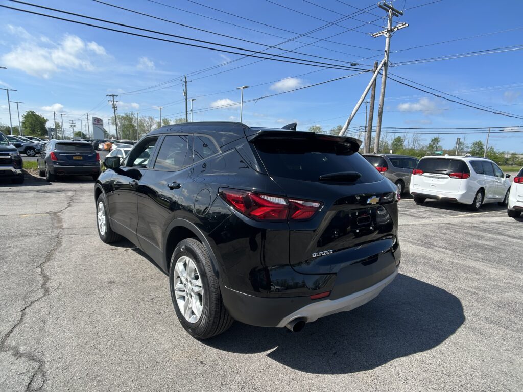 Rear three-quarter view of a black Chevrolet Blazer in a car dealership lot with other parked cars and overhead power lines on a sunny day