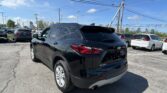 Rear three-quarter view of a black Chevrolet Blazer in a car dealership lot with other parked cars and overhead power lines on a sunny day