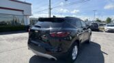 Rear three-quarter view of a black Chevrolet SUV in a car dealership lot with other cars and a white showroom on the left under a blue sky.
