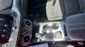 Front interior of a car showing the center console with cup holders, gear shifter, and a touchscreen dashboard near the left edge, plus fabric seats nearby.