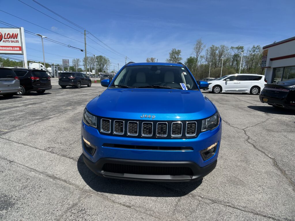 Front view of a blue Jeep SUV in a busy car dealership lot with other vehicles and a clear blue sky behind it, storefronts in the background.