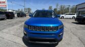 Front view of a blue Jeep SUV in a busy car dealership lot with other vehicles and a clear blue sky behind it, storefronts in the background.
