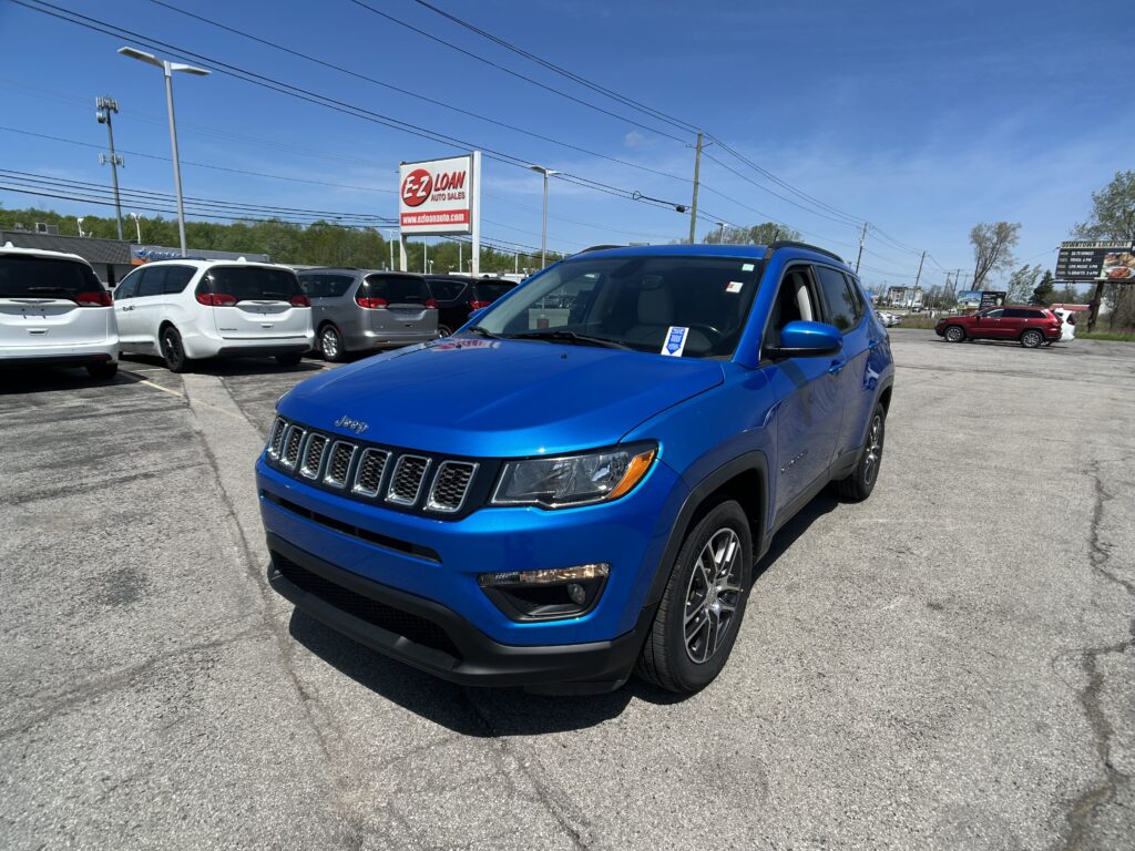 Blue Jeep SUV in the foreground at a car dealership lot with white minivans behind and an EZ Loan sign in the background.