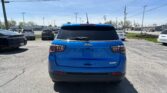 Rear view of a blue Jeep Latitude SUV parked in a crowded lot, with other cars and overhead power lines under a clear blue sky.
