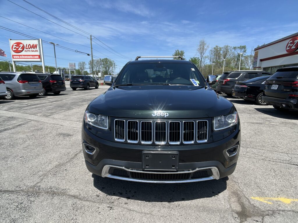 Front view of a dark Jeep SUV at a car dealership, with other cars and an E-Z Loan sign in the background under a blue sky.