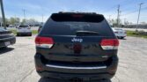 Rear view of a black Jeep SUV in a parking lot, tail lights visible, with other cars and a blue sky in the background.