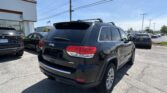Rear three-quarter view of a black Jeep SUV parked in a dealership lot with other cars and a white building in the background.