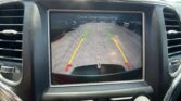 In-dash backup camera view showing a parking lot, with green and yellow trajectory lines and a center dashed line, and the message 'Check Entire Surroundings' on screen.