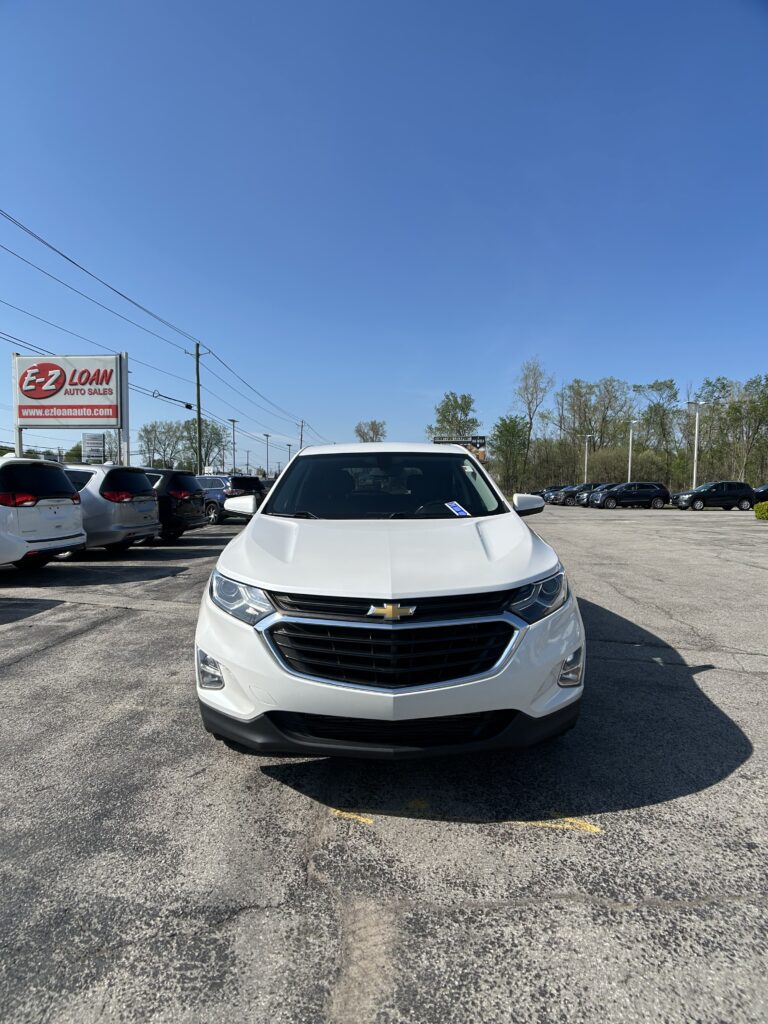 Front view of a white Chevrolet SUV in a car dealership lot with a clear blue sky above and a row of parked cars to the sides, including an E-Z LOAN Auto Sales sign on the left.