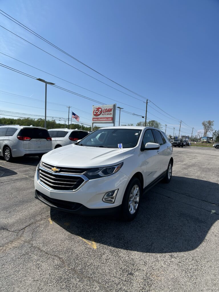 White Chevy SUV in a car dealership lot with EZ LOAN sign and an American flag in the background on a sunny day