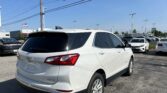 Rear three-quarter view of a white Chevrolet SUV in a car dealership lot with other cars and a clear blue sky above.