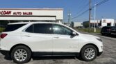 White Chevrolet Equinox SUV parked in a dealership lot with a building that reads 'AUTO SALES' and a clear blue sky in the background