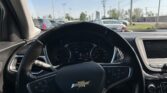 Interior view of a Chevrolet SUV from the driver’s seat, showing the steering wheel with the Chevy emblem, dashboard, and center console. Outside the windshield, a parking lot with other vehicles is visible under a clear blue sky. A sun visor sticker is on the left side of the windshield.