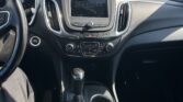 Interior view of a car dashboard focusing on the center console: touchscreen display, climate controls, gear shifter, and cup holders visible in daylight outside the windshield.