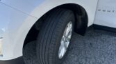 Close-up of a white SUV's front left wheel and fender with the door slightly ajar. The tire and wheel are visible against asphalt.