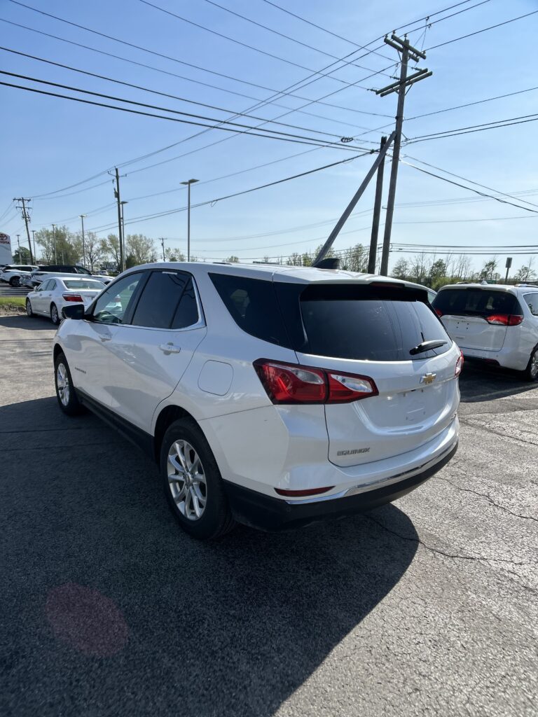 White Chevrolet Equinox SUV parked in a lot with overhead power lines against a clear blue sky, foreground-focused rear three-quarter view of the vehicle.