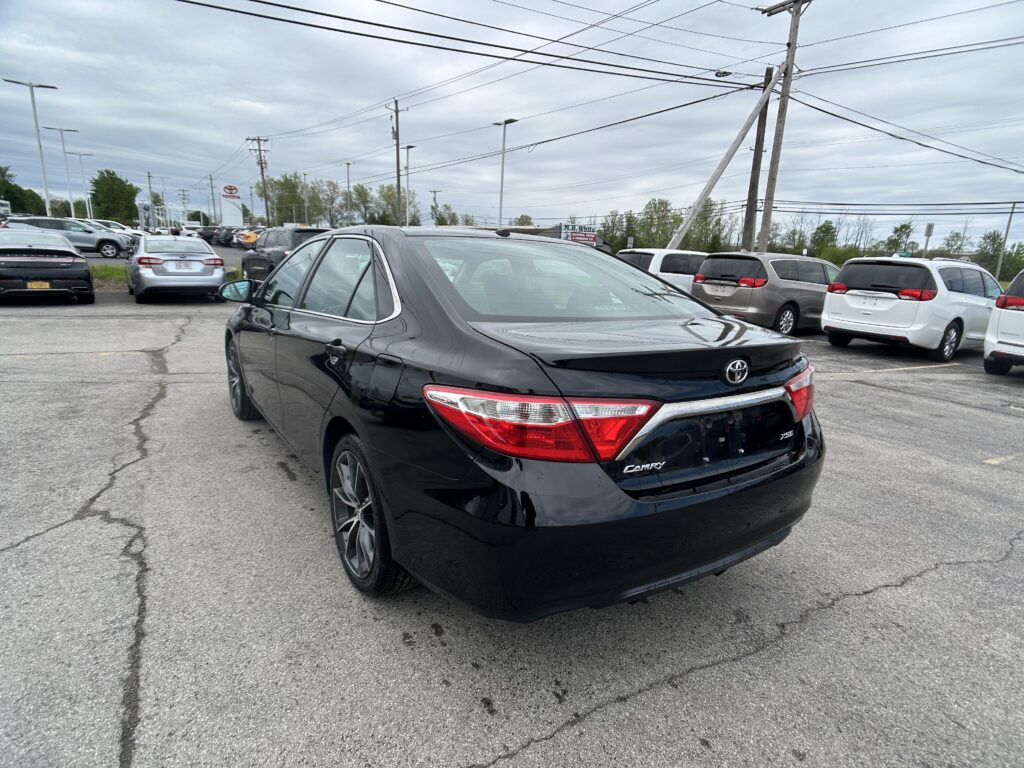 Black Toyota Camry in the foreground of a crowded car dealership lot under a cloudy sky with many parked cars and power lines overhead.