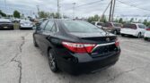 Black Toyota Camry in the foreground of a crowded car dealership lot under a cloudy sky with many parked cars and power lines overhead.