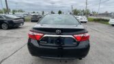 Rear view of a black Toyota Camry XSE parked in a busy car lot with other vehicles and a Toyota sign in the background.