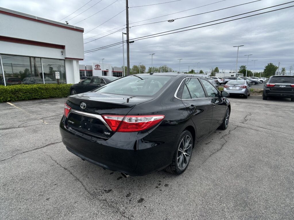 Black Toyota sedan in the foreground at a Toyota dealership lot with a showroom and many cars in the background.