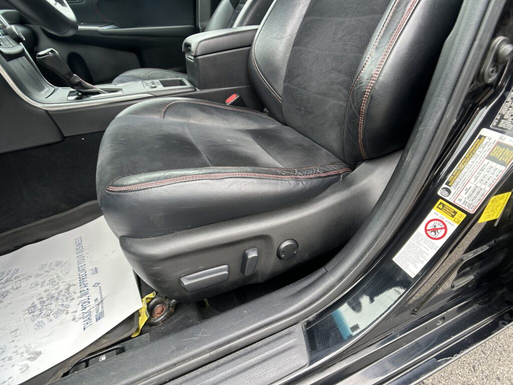 Front interior of a car showing black leather seats with red stitching, center console, and gear shifter beside the door frame with warning stickers.