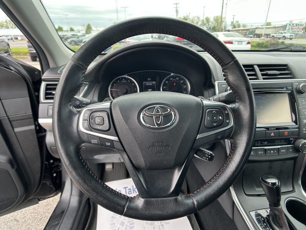 Close-up view of a black leather steering wheel from the driver's seat, with the instrument cluster and car infotainment screen in the background.