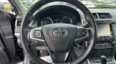 Close-up view of a black leather steering wheel from the driver's seat, with the instrument cluster and car infotainment screen in the background.