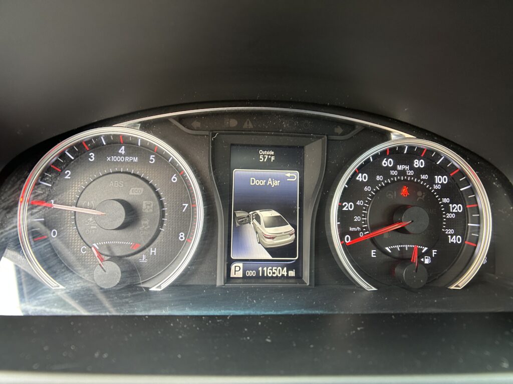 Car dashboard instrument cluster showing speedometer and tachometer with a central display reading 'Door Ajar' and vehicle icons; outside temperature 57°F, odometer 116,504 miles.