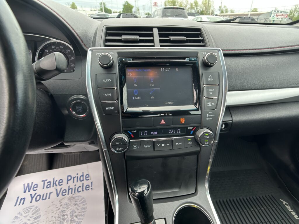 Interior view of a car dashboard: steering wheel on left, instrument cluster, central touchscreen, climate controls, and gear shifter with a paper flyer reading 'We Take Pride In Your Vehicle' visible on the left side of the seat.