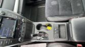 Close-up of a car’s center console: gear shifter, climate controls, touch screen, and cup holders with dark upholstery and red stitching on the seats nearby.