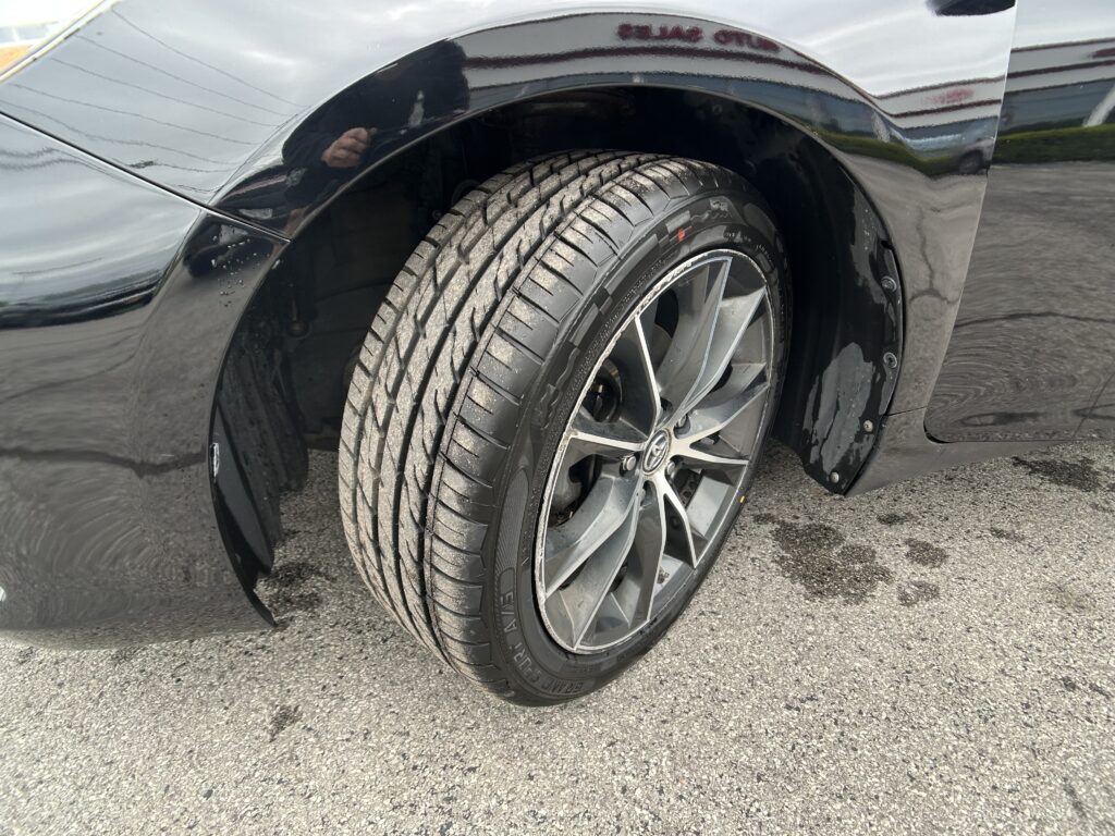 Close-up of a black car's front wheel with a treaded tire and alloy rim showing the fender above.