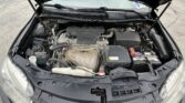 Front view of a car with the hood up, exposing the engine bay, battery on the right, and various hoses and components visible.