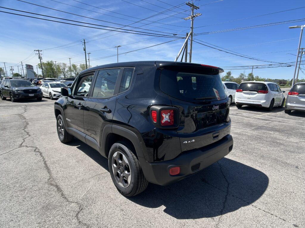 Rear view of a black Jeep Renegade in a sunny car dealership lot, with other cars and overhead power lines visible.