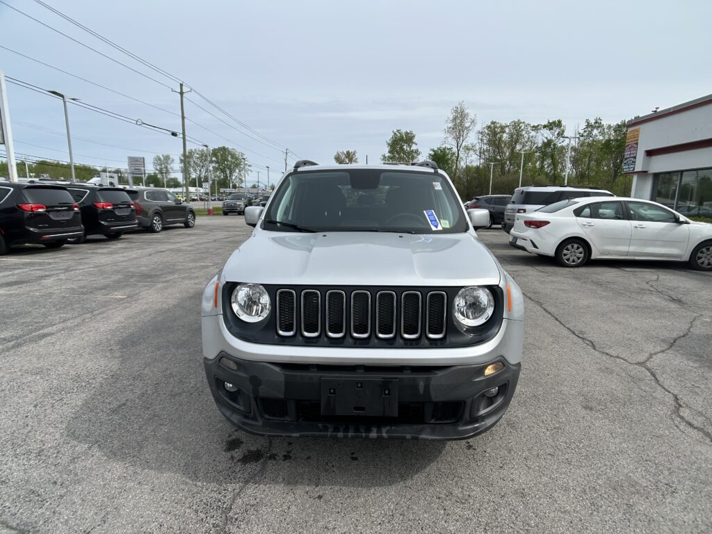 Front view of a silver Jeep SUV parked in a car dealership lot with other cars around and a building in the background.