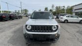 Front view of a silver Jeep SUV parked in a car dealership lot with other cars around and a building in the background.