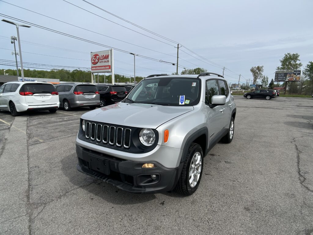 Front-right view of a silver Jeep SUV in a car dealership lot, with an E-Z LOAN Auto Sales sign and rows of minivans behind.