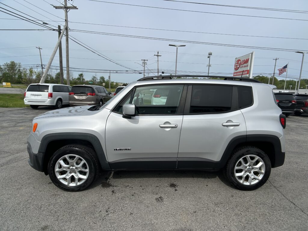 Silver Jeep Renegade SUV in a car dealership lot with other vehicles, power lines, and an American flag in the background, overcast sky.