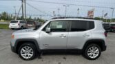 Silver Jeep Renegade SUV in a car dealership lot with other vehicles, power lines, and an American flag in the background, overcast sky.