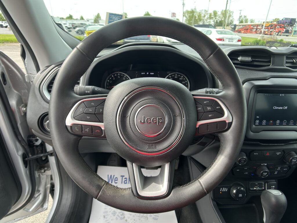 Driver's seat view showing a black Jeep steering wheel, dashboard, and center console with a bright car lot seen through the windshield edge.