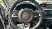 Driver's seat view showing a black Jeep steering wheel, dashboard, and center console with a bright car lot seen through the windshield edge.