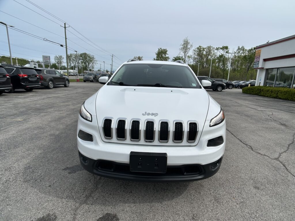 Front view of a white Jeep SUV in a car dealership parking lot with other vehicles in the background, sky overhead.