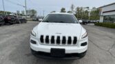 Front view of a white Jeep SUV in a car dealership parking lot with other vehicles in the background, sky overhead.