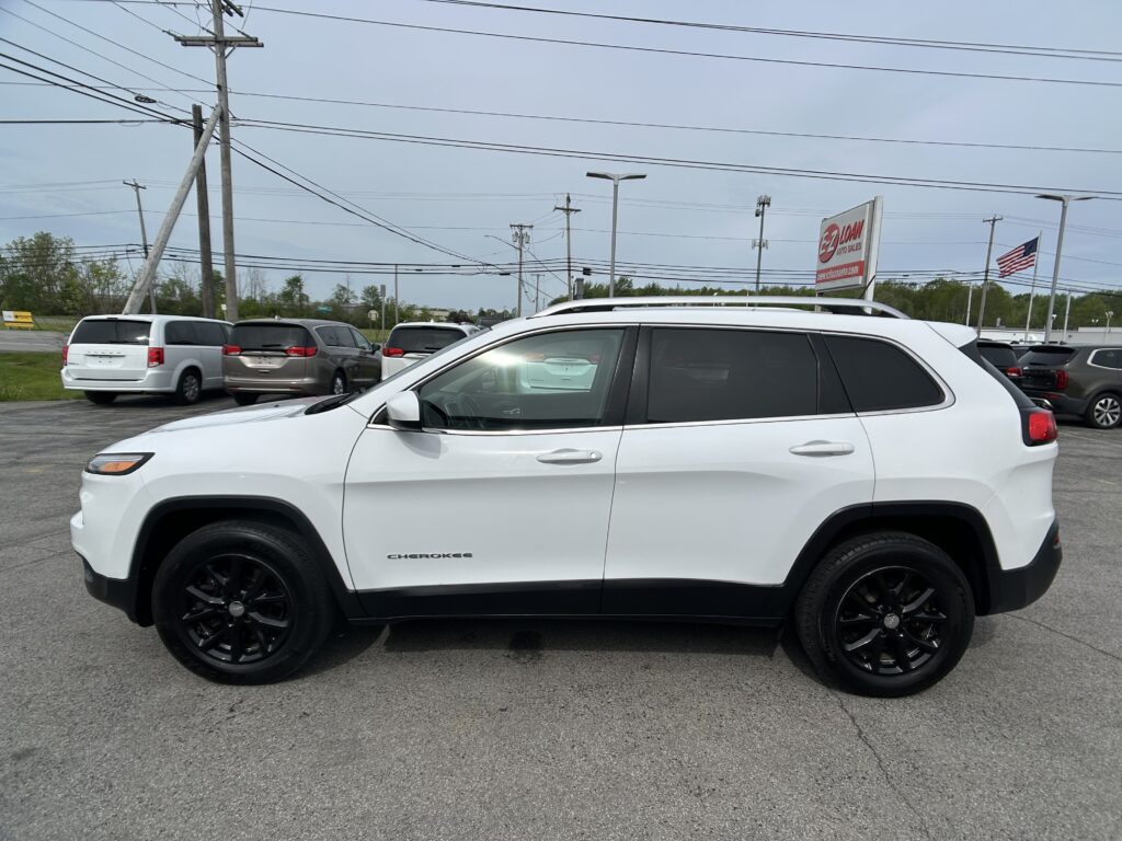 White Jeep Cherokee in a car dealership lot with several minivans in the background and power lines overhead.