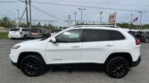 White Jeep Cherokee in a car dealership lot with several minivans in the background and power lines overhead.