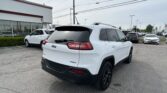 White Jeep SUV viewed from the rear in a car dealership lot, with other cars and a showroom building in the background