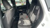 Back seat of a dark fabric car interior with three seats, headrests, and visible seat belts; parking lot seen through the rear window.