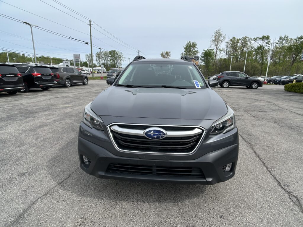 Front view of a gray Subaru SUV centered in a busy car dealership lot with other vehicles parked on both sides under a cloudy sky.