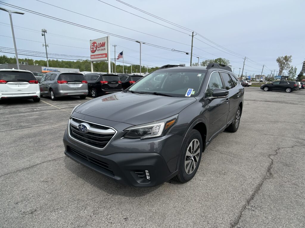 Front-right corner of a gray Subaru SUV in a car dealership lot with multiple vehicles in the background and power lines overhead, under a blue sky.